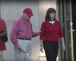 Woman guiding a man wearing a baseball cap. 